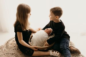 Two children sit on a round rug while playing with a baby in a minimalist white room.