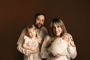 A family of four wearing cream-colored outfits poses together against a dark brown studio backdrop.