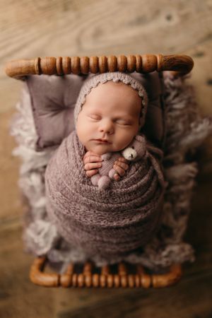 A sleeping newborn baby swaddled in soft gray knit fabric rests peacefully on a wooden surface.