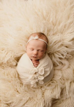 A sleeping newborn swaddled in white fabric lies peacefully on a fluffy cream-colored fur backdrop.