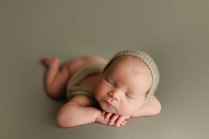 Sleeping newborn wearing knit bonnet rests peacefully on olive green backdrop with crossed arms.