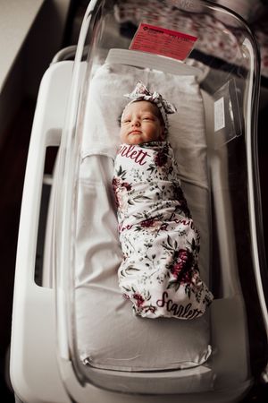 Newborn baby swaddled in floral print blanket resting inside clear hospital bassinet.