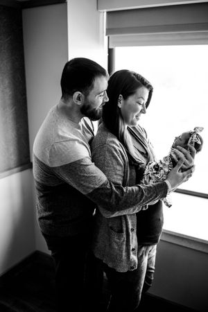 Black and white photo of couple gazing at newborn baby near window in hospital room.