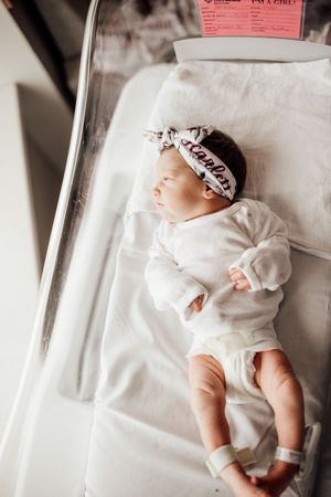 A newborn baby wearing white clothing lies peacefully on white bedding in a hospital bassinet.