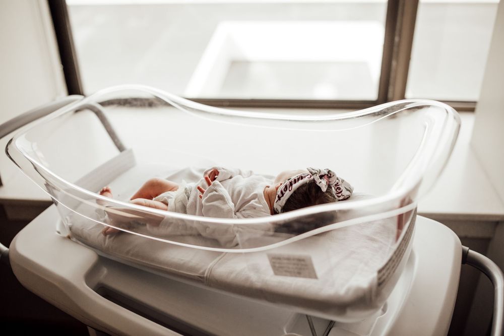 Newborn baby sleeps peacefully in a clear hospital bassinet near a window.