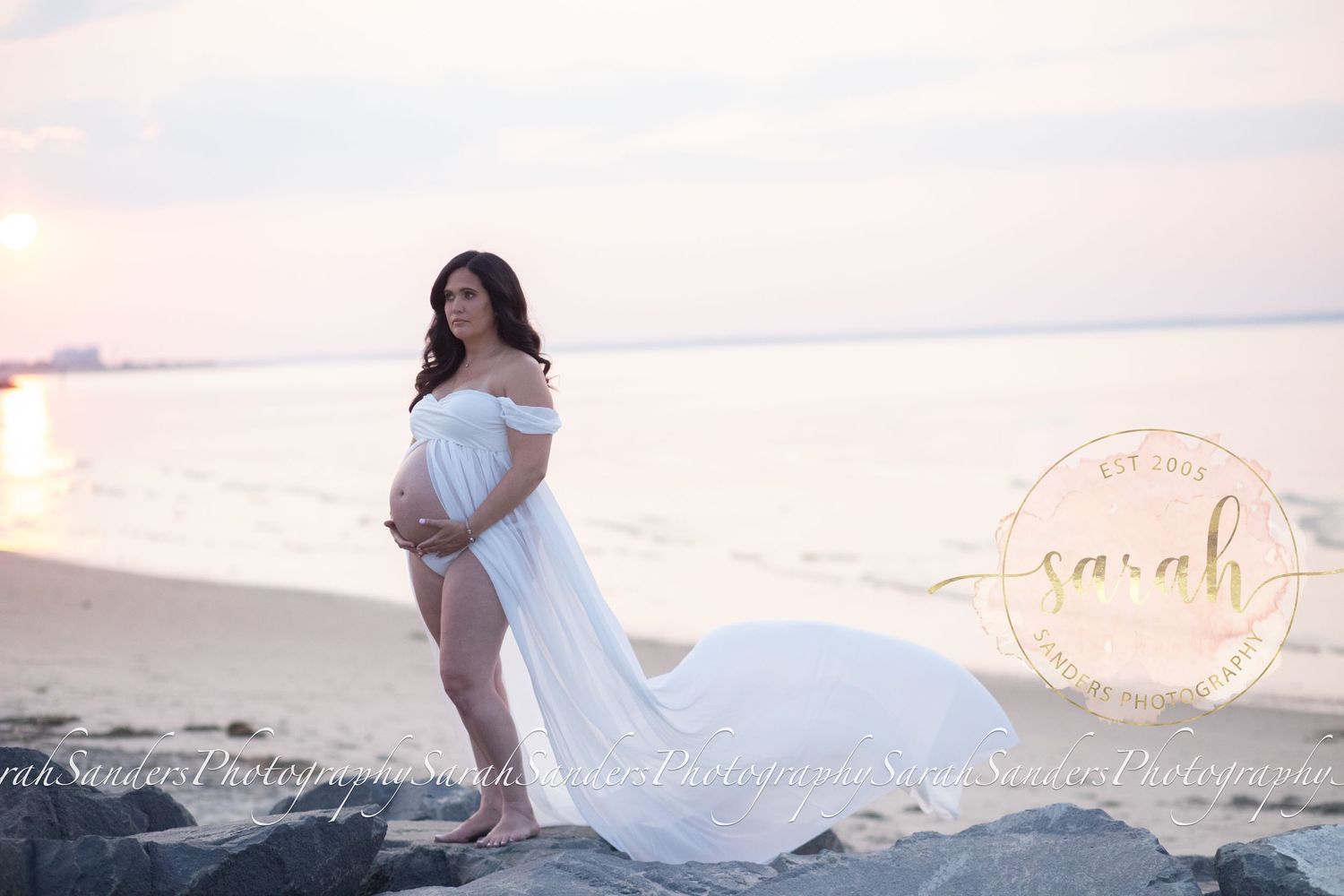 Pregnant woman standing posed with flowing sheer in the wind, on rock jetty at beach during sunset