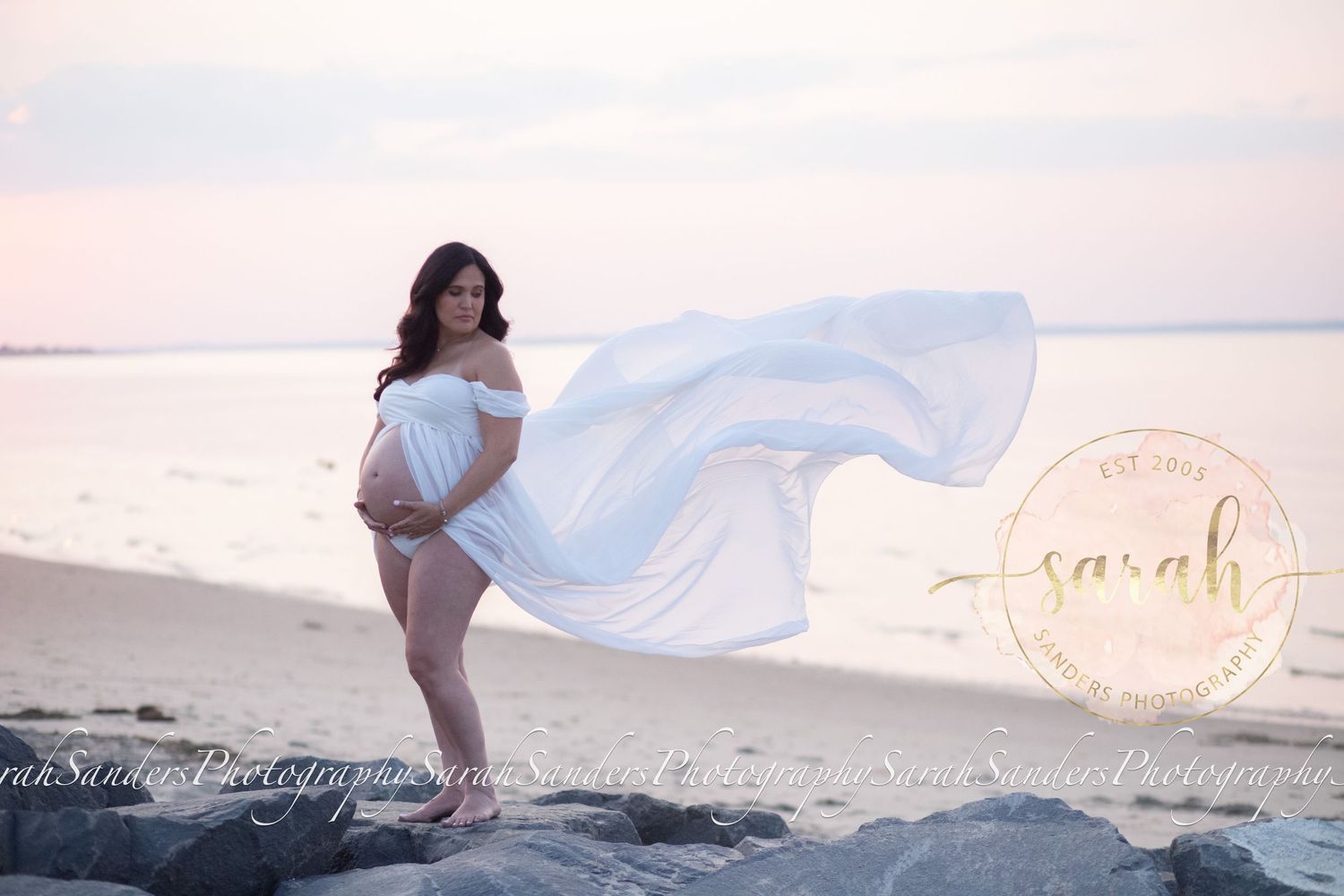 Pregnant woman standing posed with flowing sheer in the wind, on rock jetty at beach during sunset