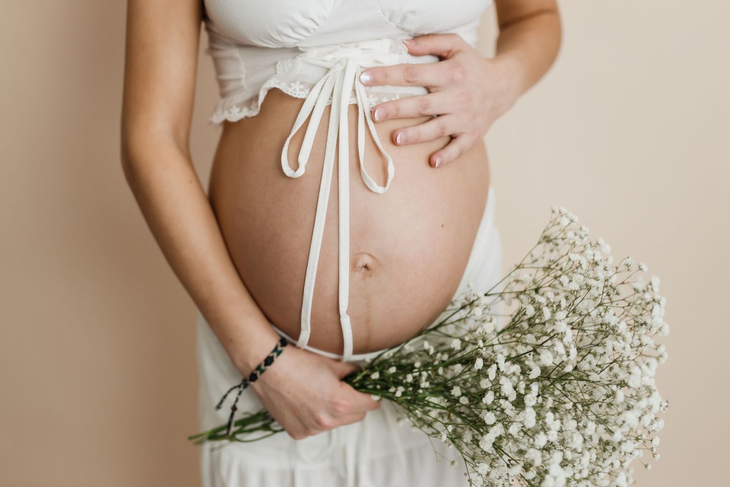 A pregnant belly with white garments and a delicate bouquet of baby's breath flowers held against a beige background.