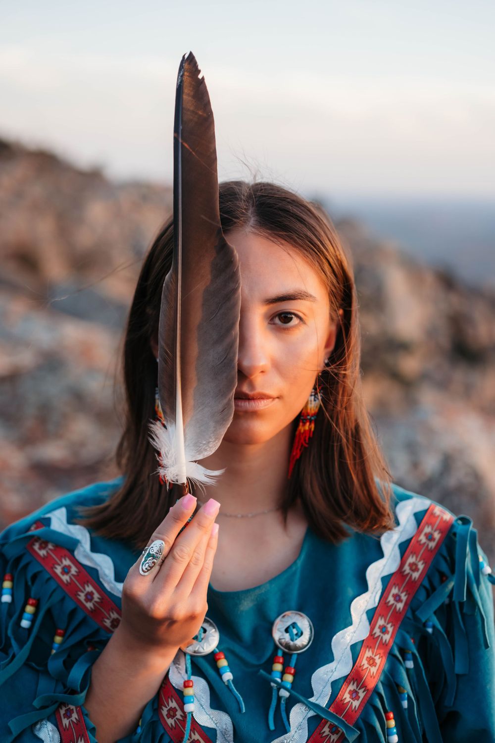 Portrait with traditional Native American feather and turquoise dress against rocky landscape.
