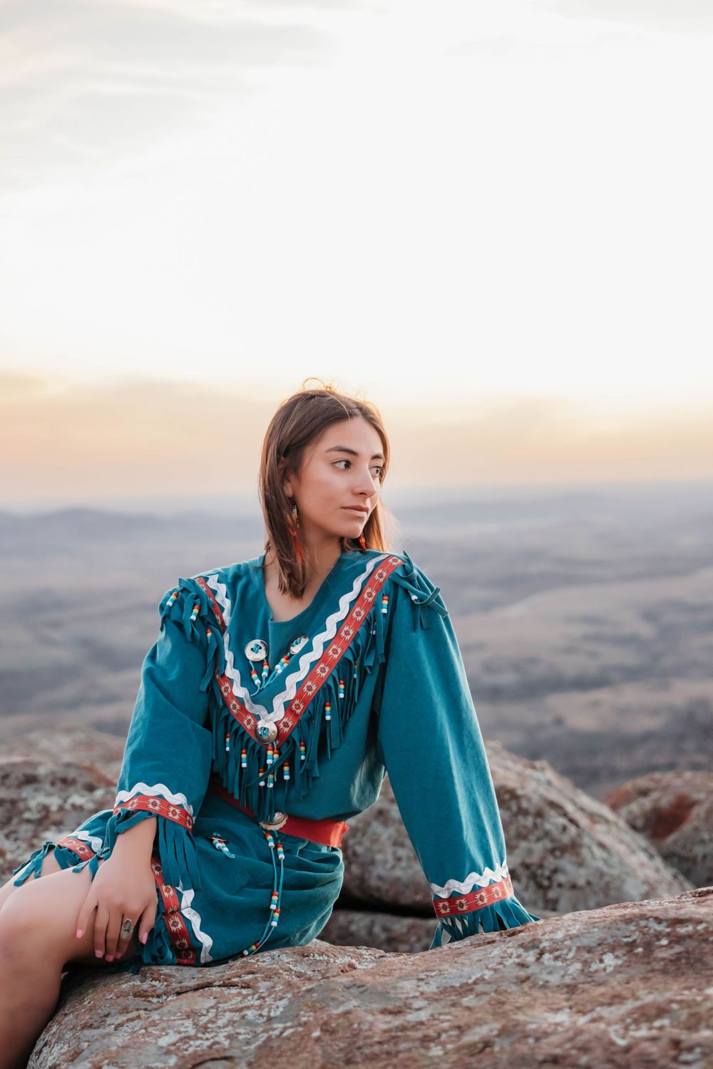 Person in decorative turquoise dress sitting on rocky overlook at sunset.