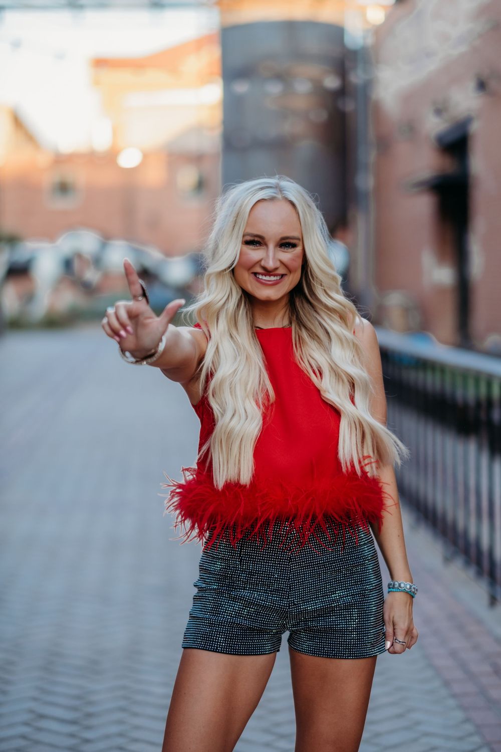 Woman wearing red feathered top and patterned shorts poses on cobblestone street.