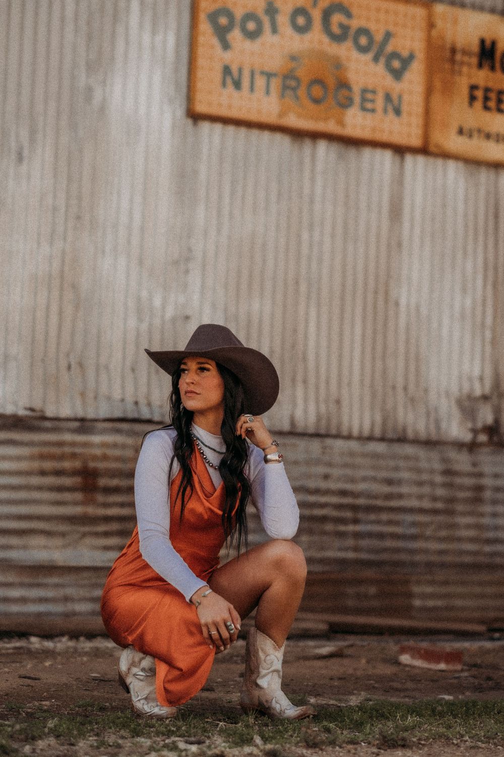 Fashion portrait in western attire crouching against industrial metal wall backdrop.