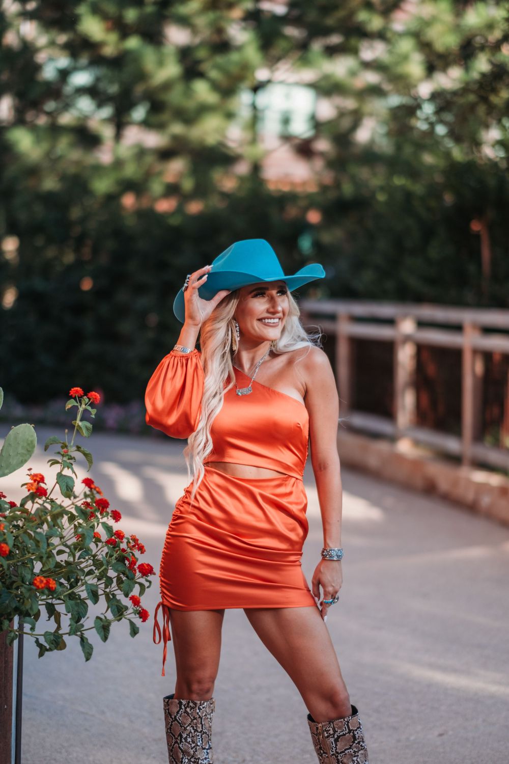 Woman in orange dress and blue cowboy hat poses outdoors with flowers.