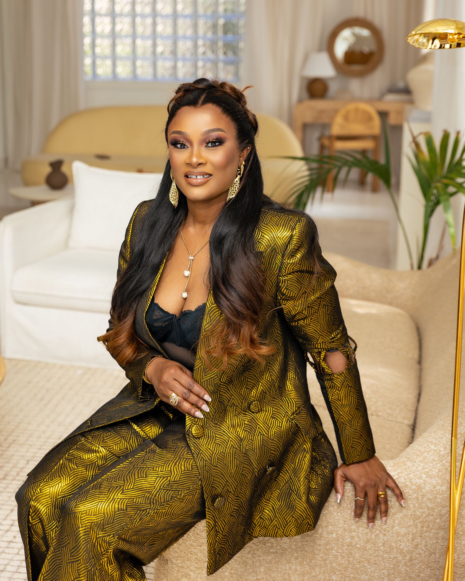 A well-dressed person wearing a shimmering gold metallic pantsuit poses on a cream colored couch in an elegant living room.