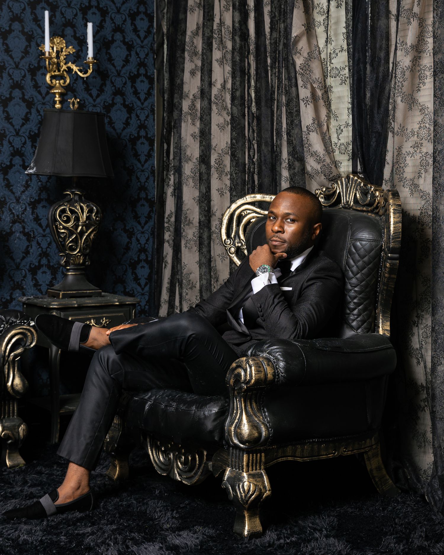Man in black suit lounges elegantly on ornate vintage chair against dark damask wallpaper and drapery with baroque decor.