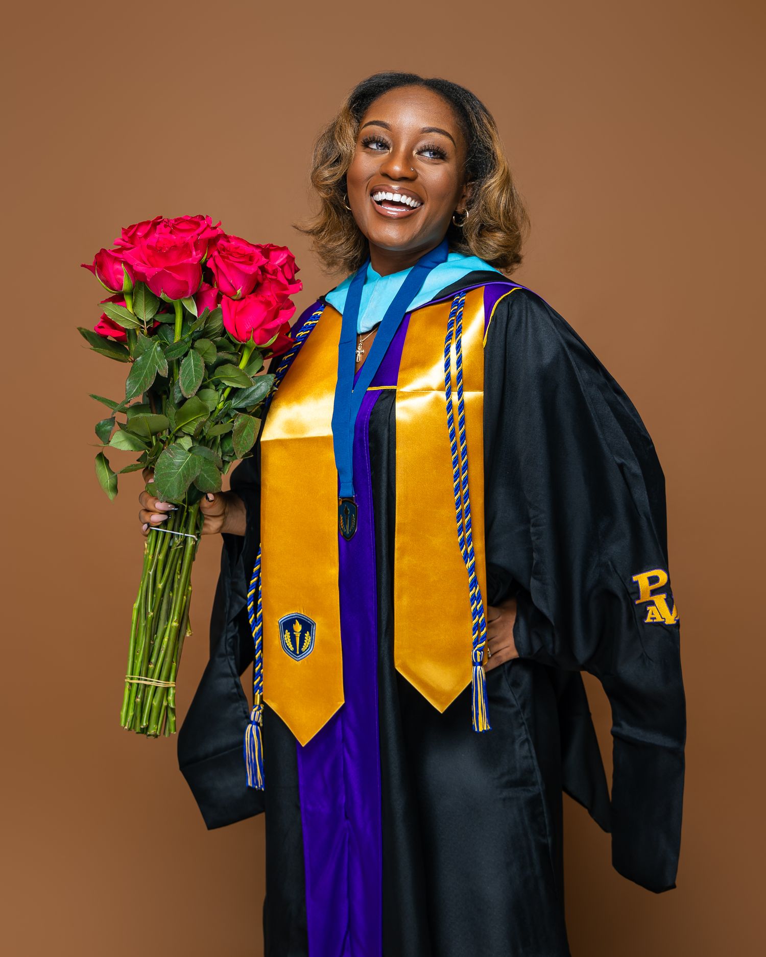 Graduate in academic regalia wearing a gold stole and honor cords holding bright red roses during commencement.