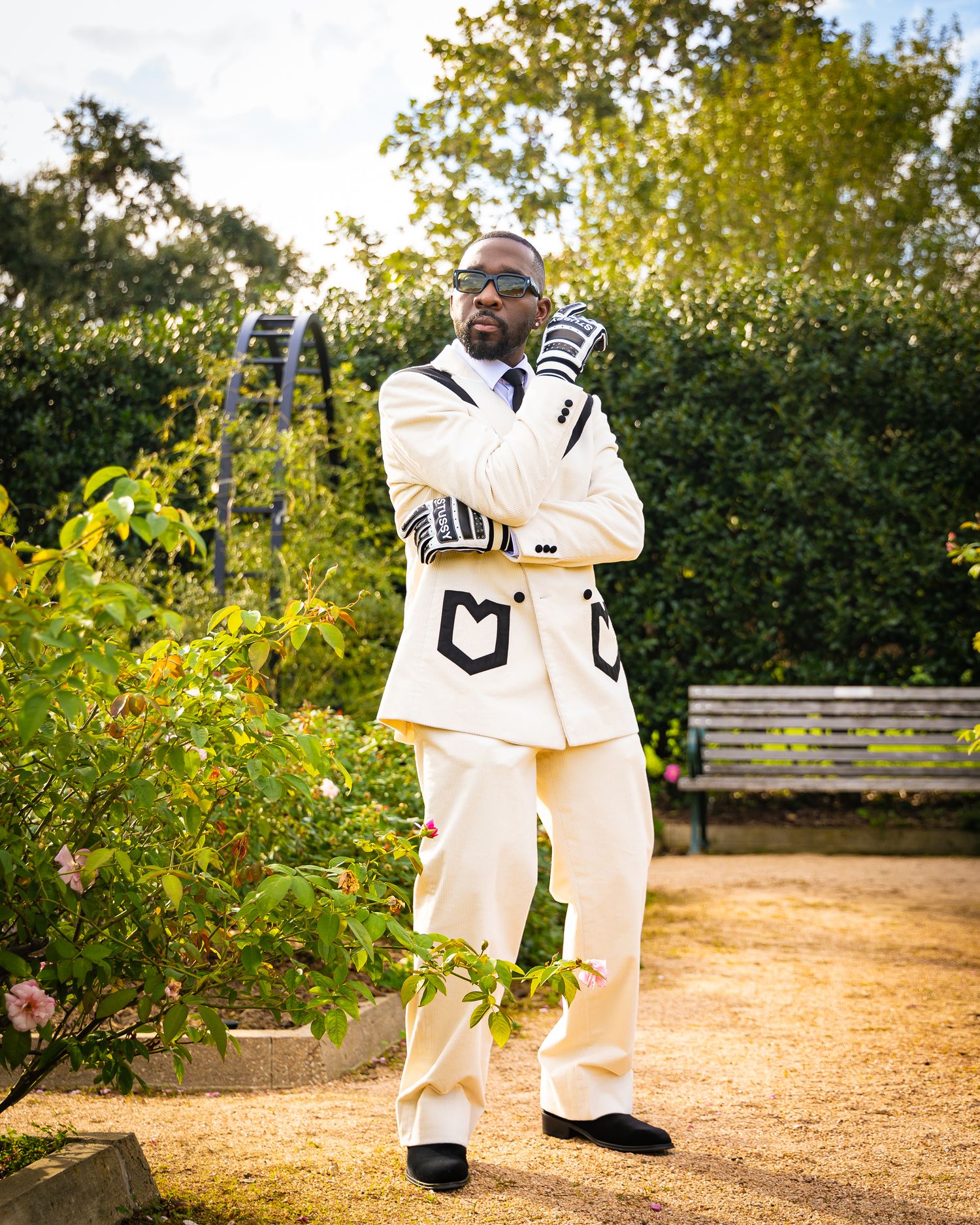 A person in a crisp white outfit with gloves poses confidently on a garden path at sunset with lush greenery in the background.