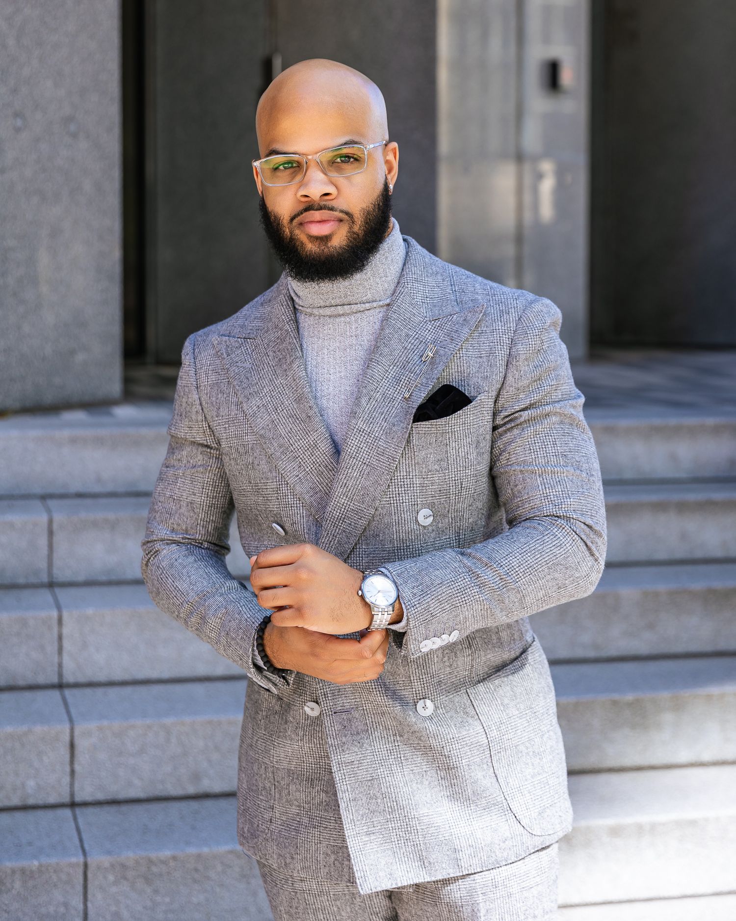 A well-dressed person in a grey double-breasted suit and turtleneck sweater poses confidently against a concrete building.