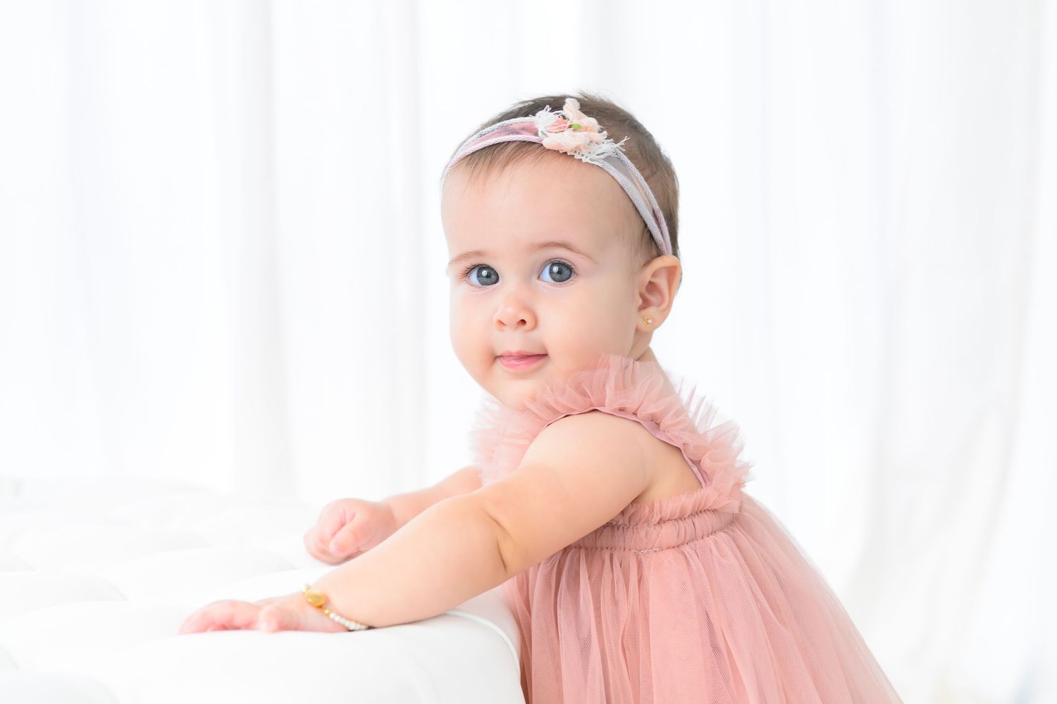 Small baby in pink dress with lace trim and sparkly headband sits elegantly against white background.