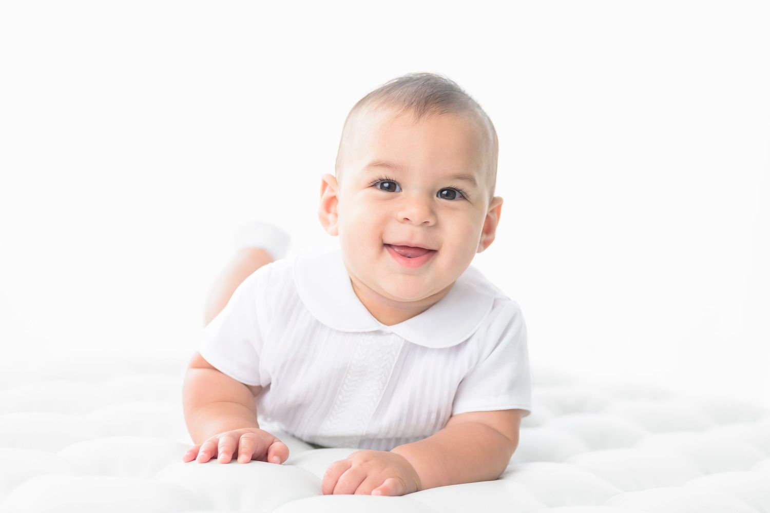 A smiling baby in white clothing lies on clean white bedding and looks at the camera.