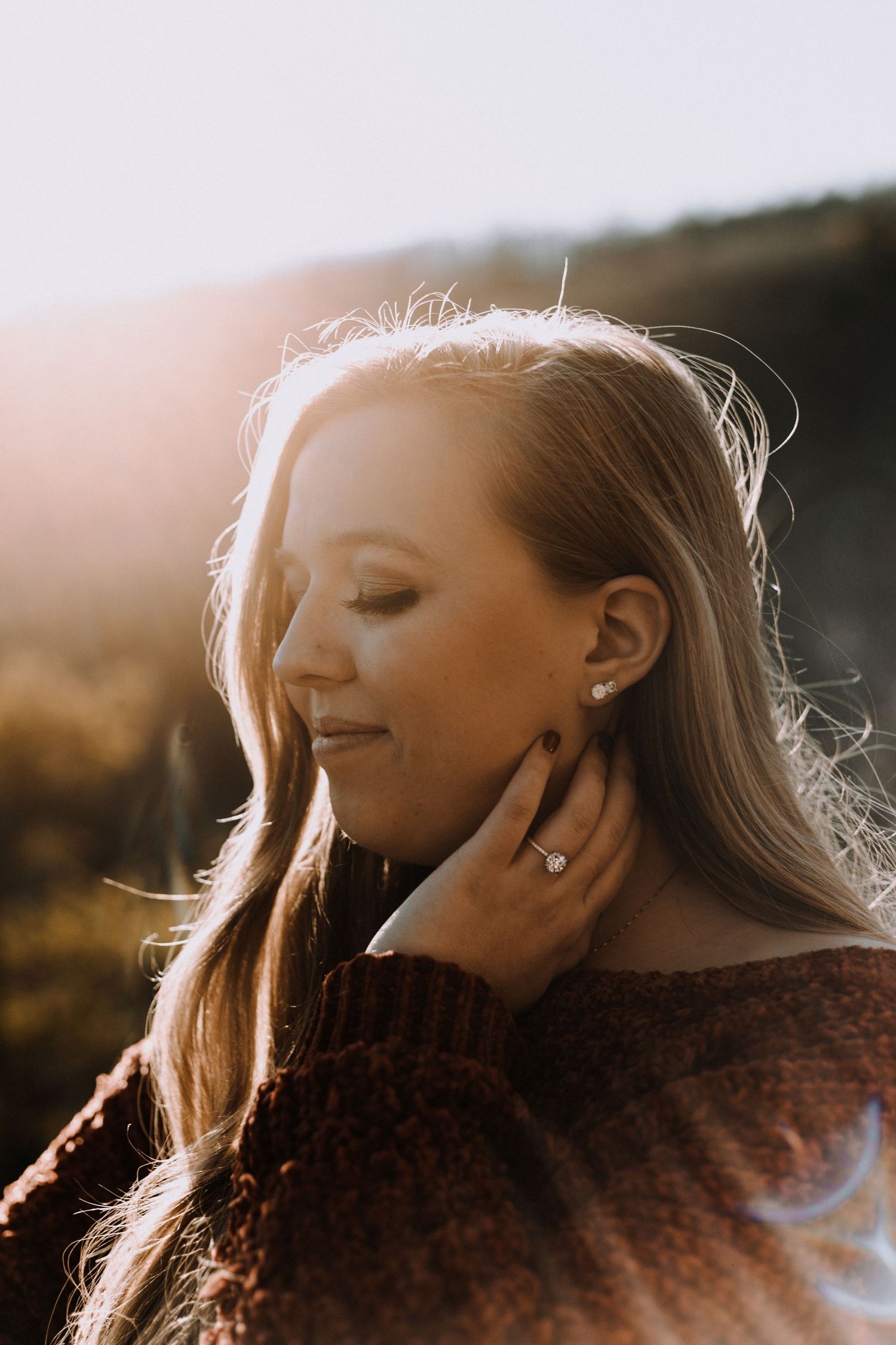 A dreamy golden-hour portrait of a woman in a burgundy sweater showing off her engagement ring.