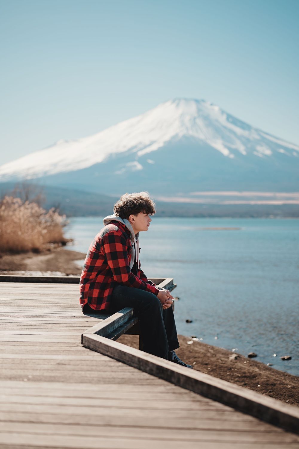 Person in red plaid shirt sits on wooden dock overlooking serene lake with snow-capped Mount Fuji in the background in Japan.