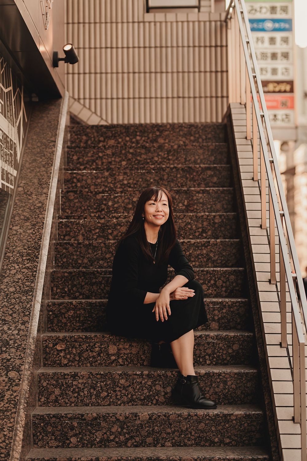 Figure in a black outfit sits on speckled concrete stairs with tiled walls and handrails in an urban setting.