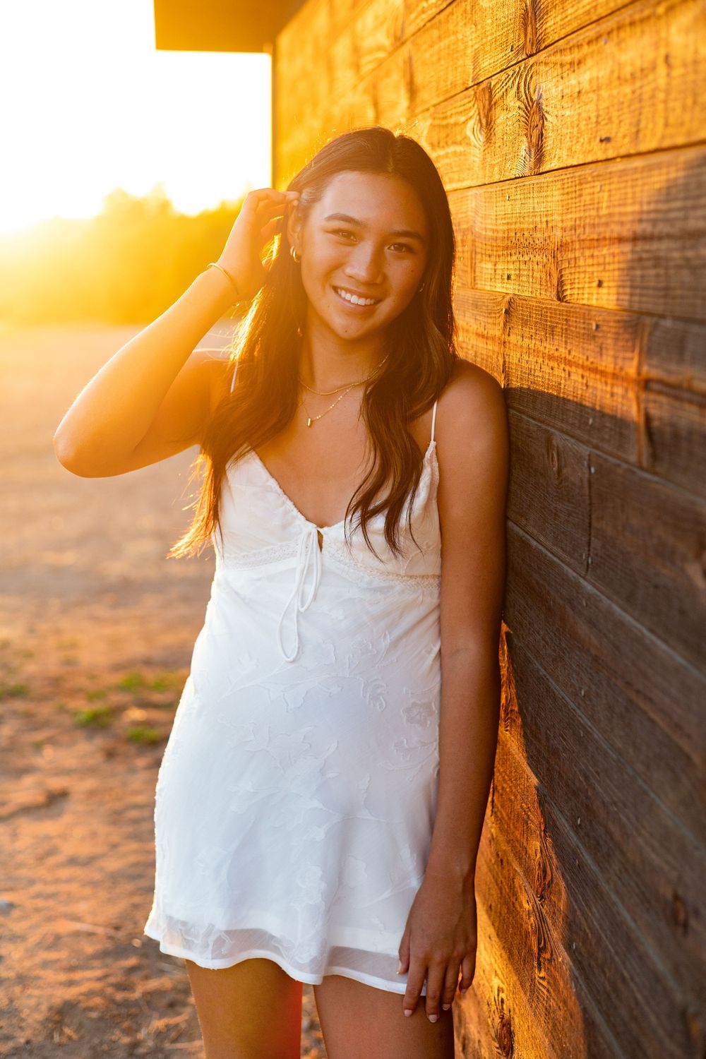Young woman in white sundress smiling against rustic wood wall in golden sunset light.