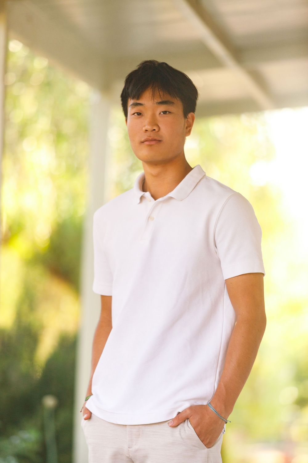 Young Asian man in white polo shirt standing outdoors on a porch with green foliage in the background.