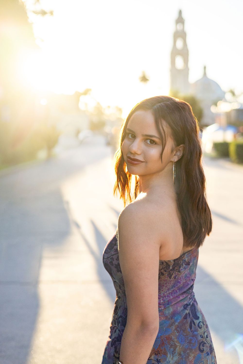 Young woman in colorful strapless dress poses on sunlit urban walkway with historic tower in background.