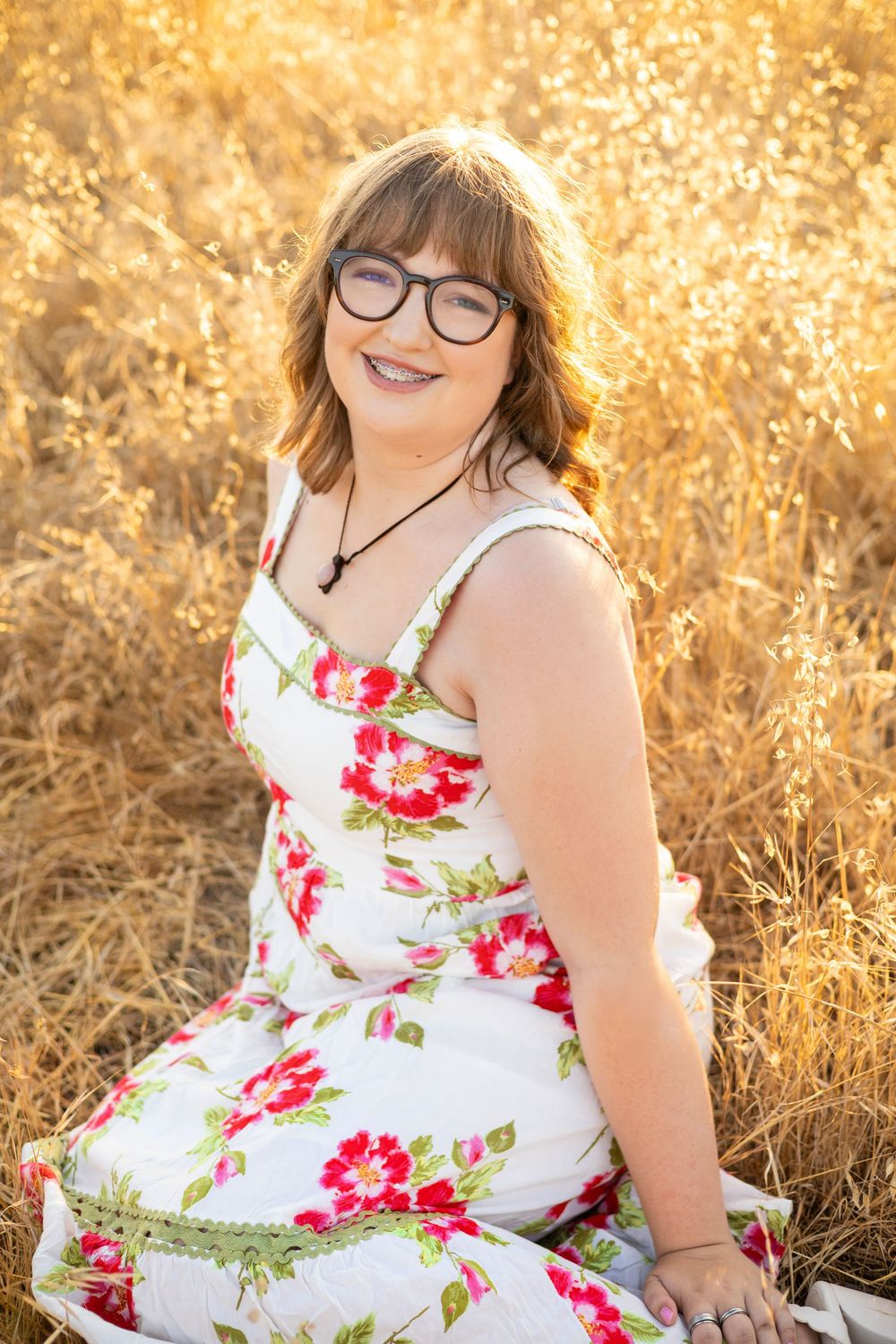 Smiling woman with glasses in floral sundress sits in golden wheat field during warm sunset light.