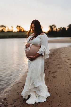 A pregnant figure in a flowing white dress stands on a beach at sunset, cradling their belly in silhouette.