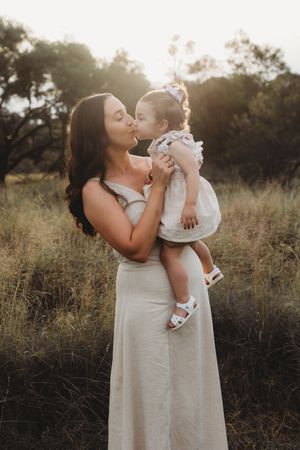A tender moment captured at sunset in a field as parent and child share an affectionate kiss in matching light-colored outfits.