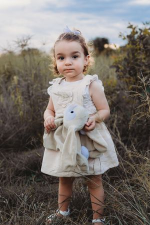 A small child in a cream-colored dress holds a stuffed animal while standing in a natural outdoor setting.