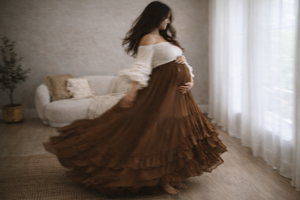 Woman in a flowing brown dress twirling in a sunlit room with white curtains.