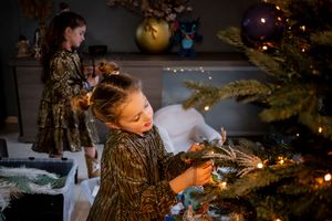 Two girls in gold dresses decorating a Christmas tree with lights and ornaments in a cozy holiday setting.