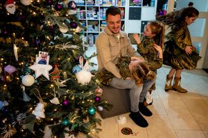Family celebrating Christmas near a decorated tree, father sitting with young daughters in festive outfits indoors.