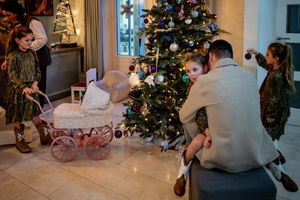 Family decorating a Christmas tree together in a festive living room with gifts and a vintage doll pram nearby.
