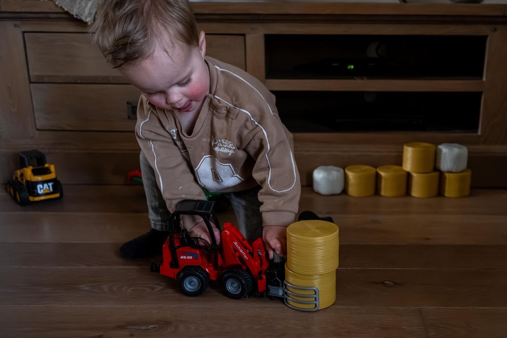 A child plays with yellow and blue toy trucks and blocks on a hardwood floor in a dimly lit room.