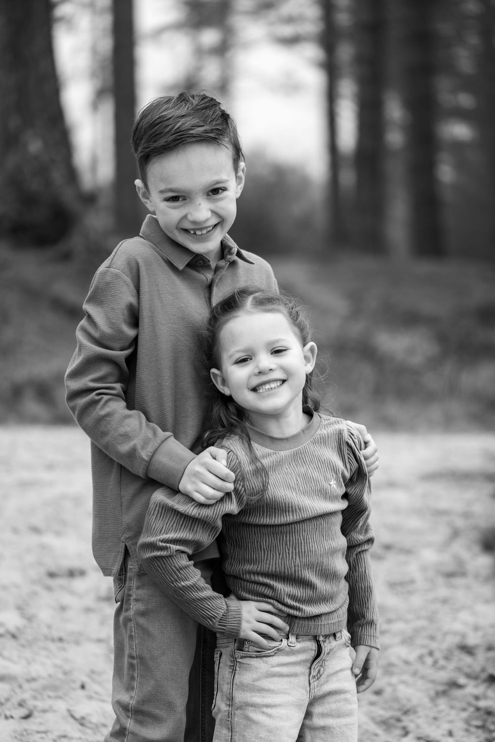 Black and white candid photos show two children playing and laughing together outdoors in a forest setting.