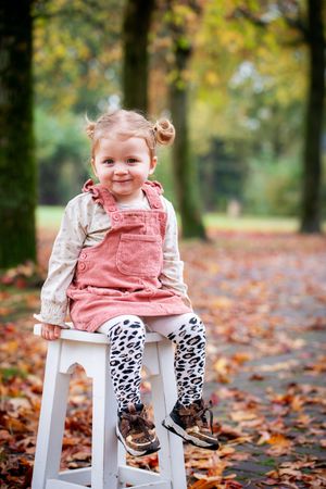 Small child in pink corduroy overalls and animal print tights sits on white stool surrounded by autumn leaves in park.