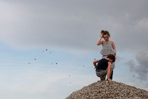 A person sits atop a rocky hill silhouetted against a cloudy sky at sunset.