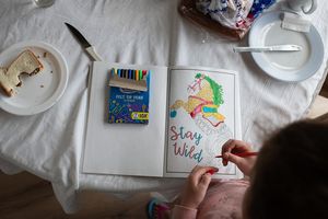 Someone draws colorful illustrations on white paper at a table with art supplies.