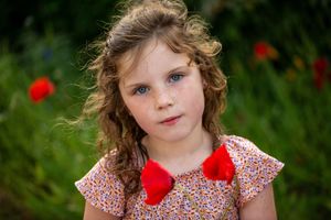 A child in a floral dress with a red bow sits in a garden with red poppies in the background.