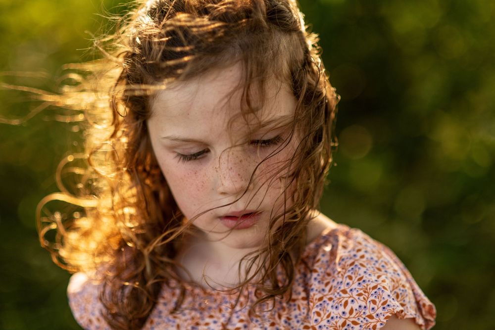 A young child in a floral dress stands outdoors with sunlit hair blowing in the breeze during golden hour.