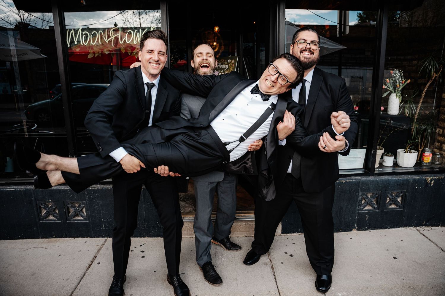 Four men in black suits laughing and lifting one man outside a restaurant.