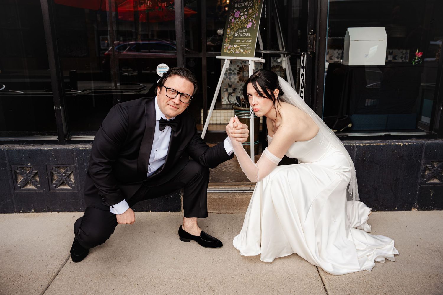 Bride and groom arm wrestling playfully while seated on sidewalk outside venue.