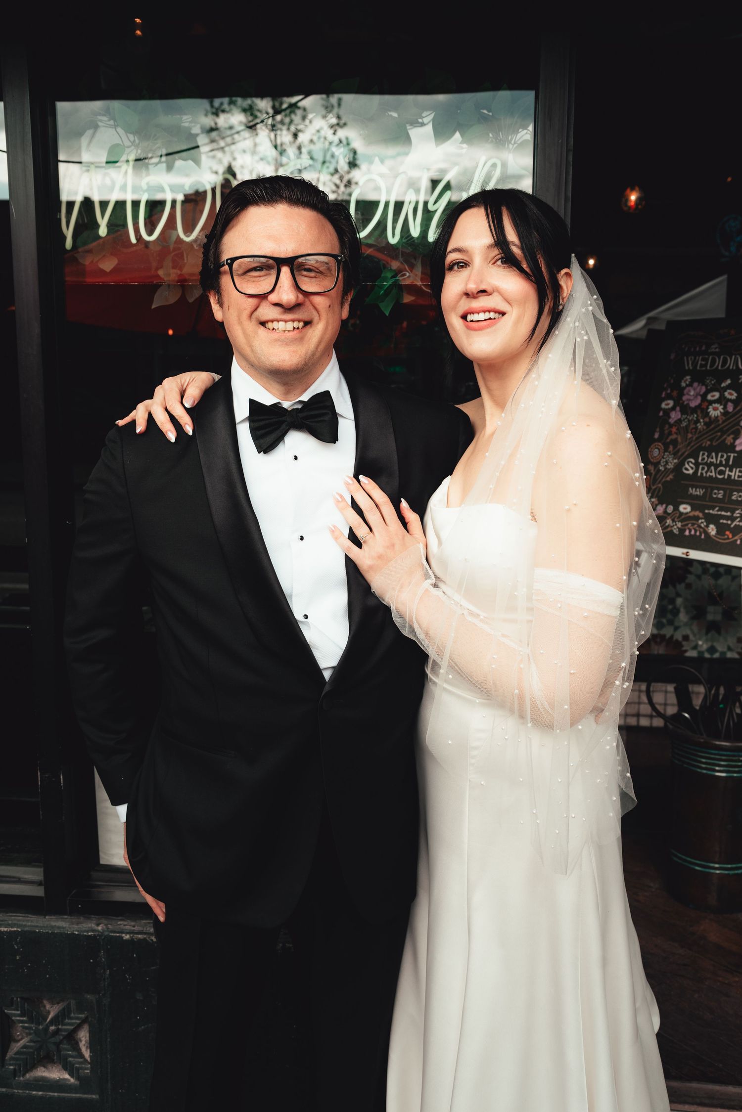 Smiling bride and groom posing together outside wedding venue with neon sign backdrop.