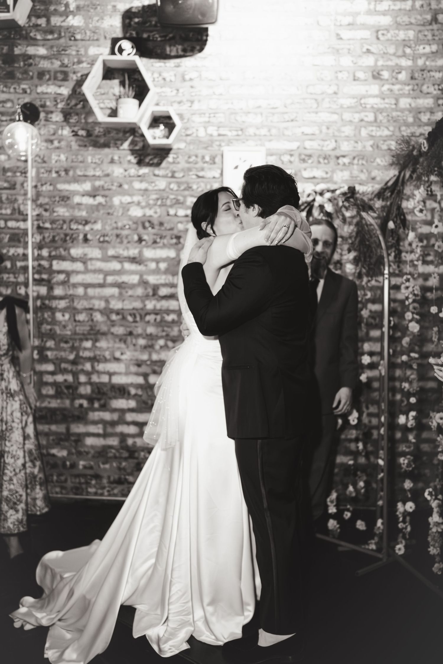 Black and white photo of bride and groom sharing first kiss at wedding ceremony near brick wall.