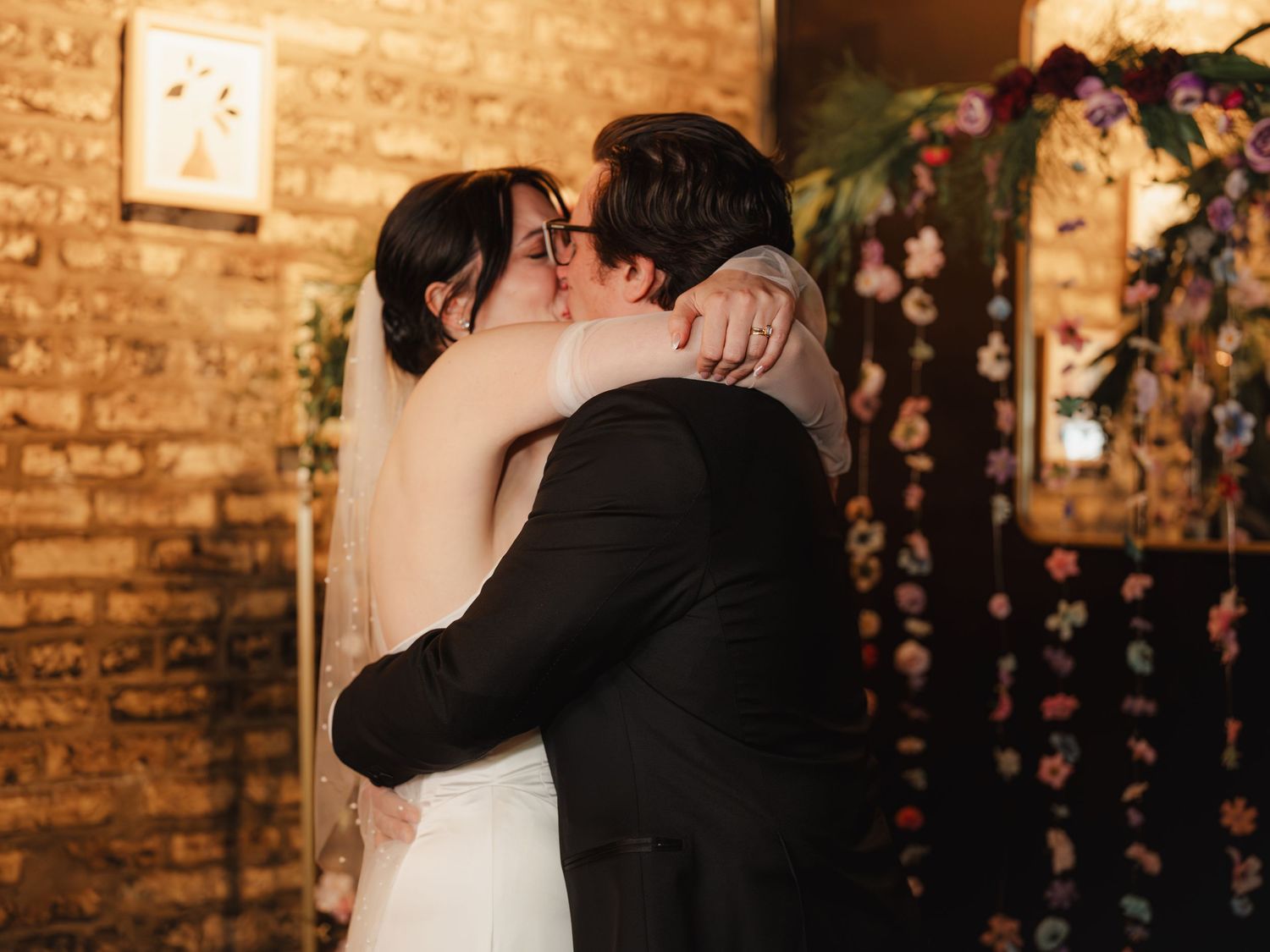 Bride and groom share their first kiss at a rustic brick-walled wedding venue with floral arch backdrop.